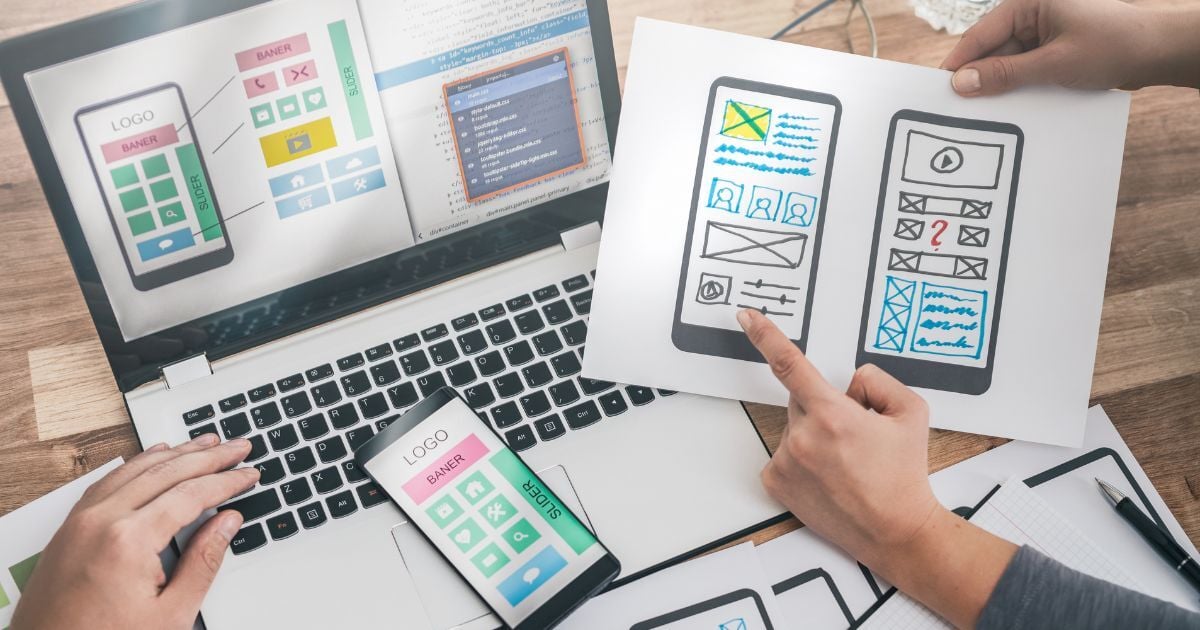 Hands reviewing mobile website wireframes and UI mockups displayed on paper, smartphone, and laptop during a website design and development planning session